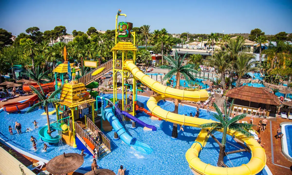 Un parque acuático con toboganes amarillos y azules, palmeras y gente disfrutando. ¡Un lugar ideal para camping con toboganes en España!