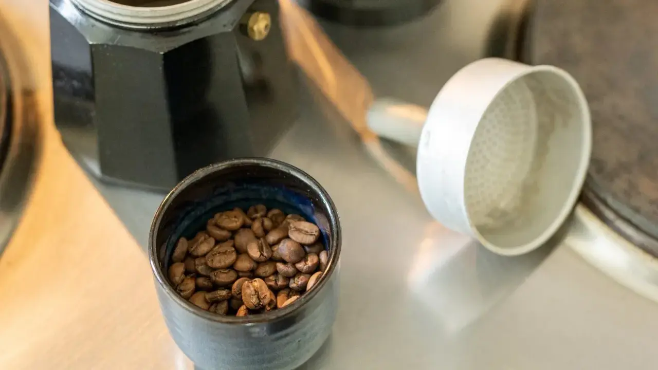 Kaffeebohnen in einer Schale neben einer Bialetti-Kaffeemaschine. Die Frage "bialetti wie viel kaffee" wird durch die Zubereitung beantwortet.