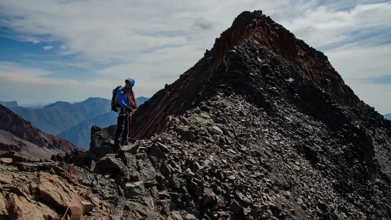 Alpinista na grani, zdobywający najwyższy szczyt Gruzji. W tle skaliste pasmo górskie i błękitne niebo.