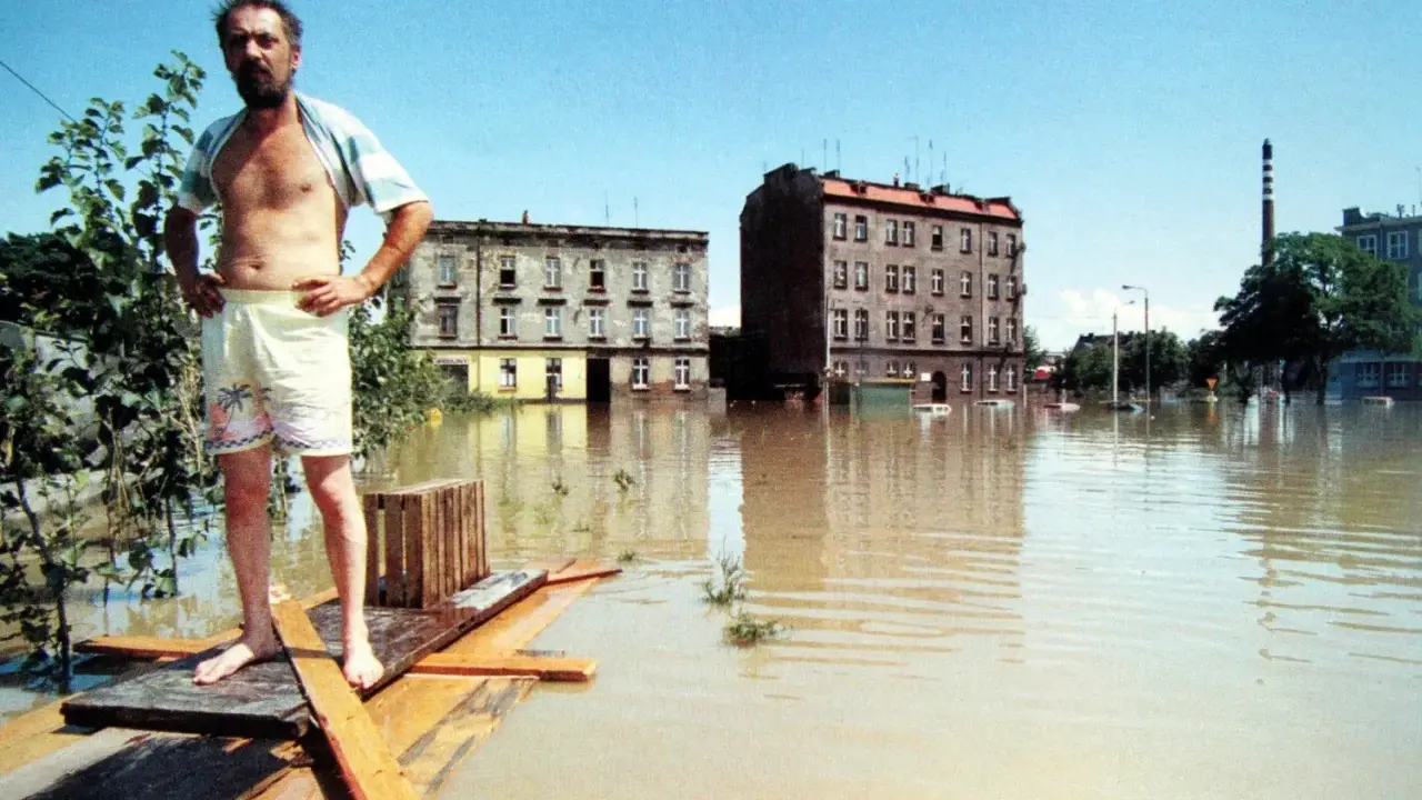 Mężczyzna na prowizorycznej tratwie w zalanej wodą ulicy. Budynki i samochody toną w brązowej wodzie. To obraz powodzi 1997.