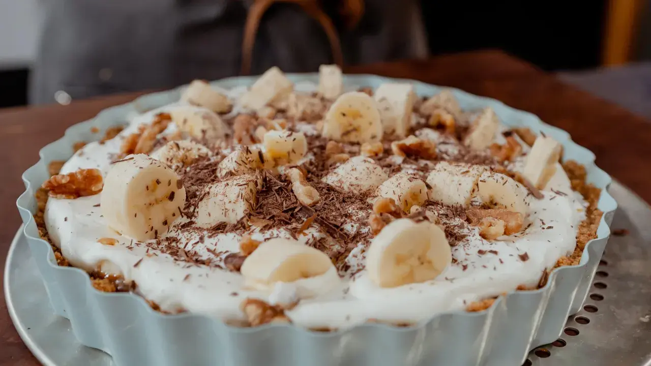 Tarte banane caramel onctueuse, garnie de rondelles de banane, noix et copeaux de chocolat. Un délice gourmand.