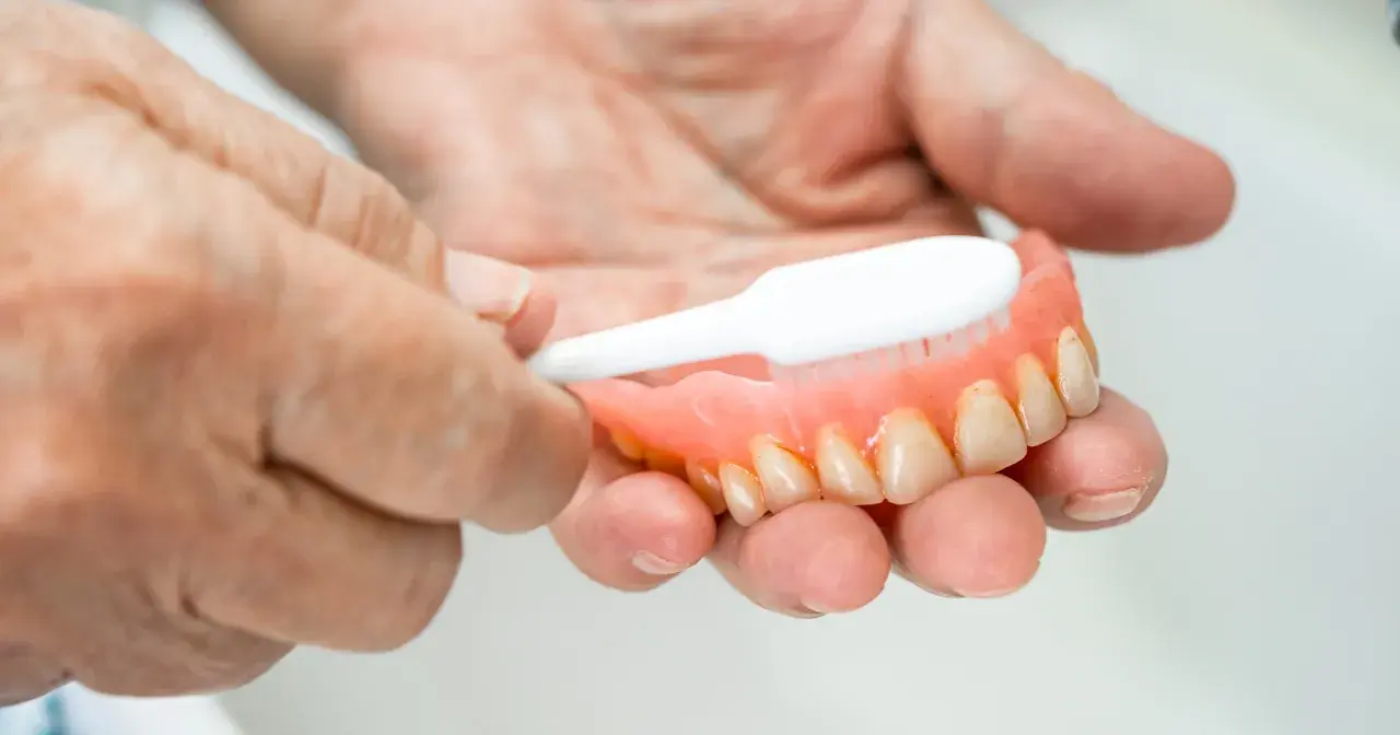 Dłonie czyszczą protezę zębową szczoteczką, pokazując jak usunąć kamień z plastiku.