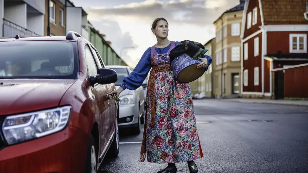 Lina Odell, klädd i folkdräkt, öppnar bildörren med en korg i famnen.