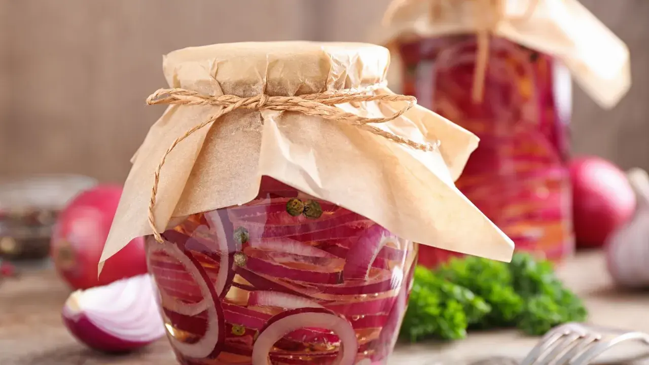Zwei Gläser mit eingelegten Zwiebeln, verziert mit Papier und Schnur. Daneben liegen frische Zwiebeln und ein Gabel.