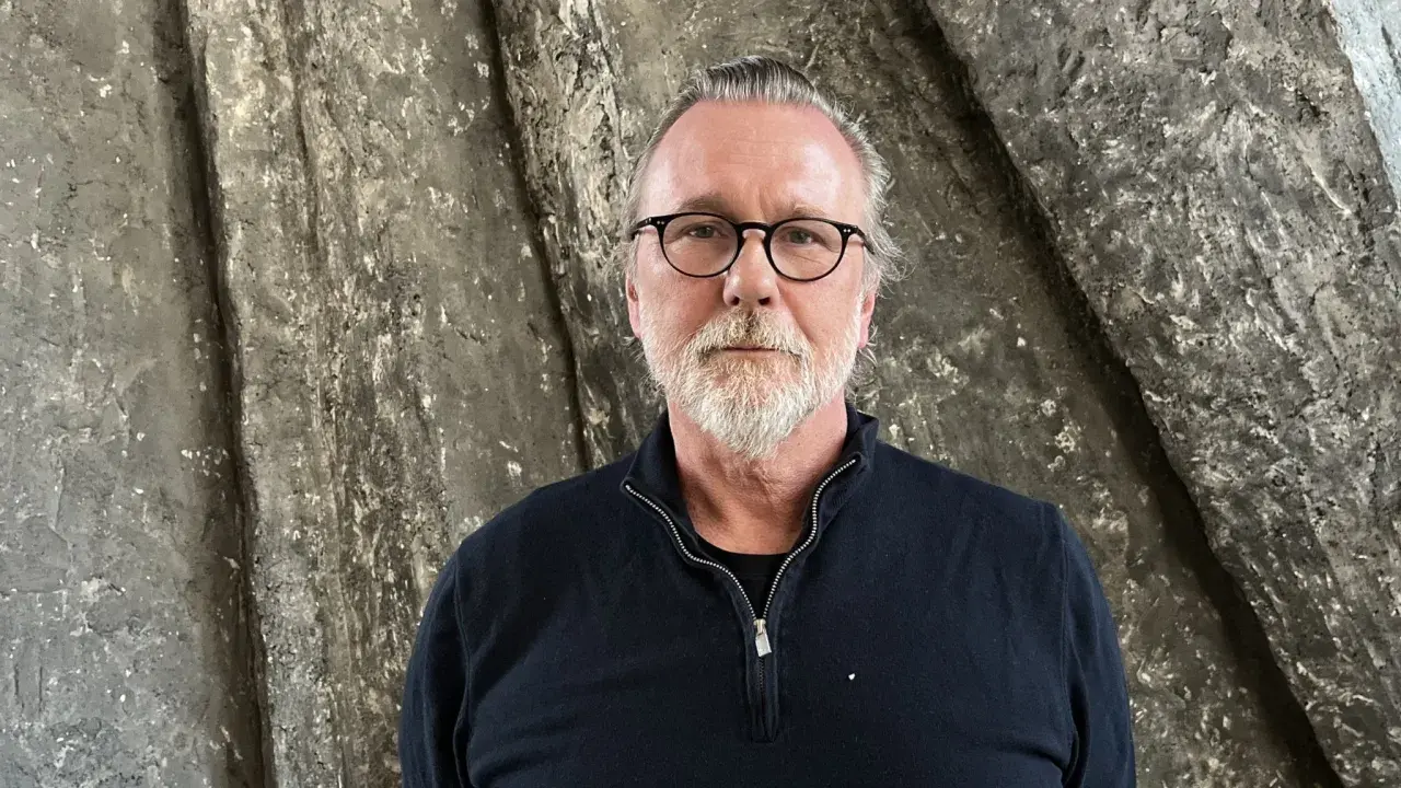 Thomas Olsson, en man med glasögon och grått skägg, står framför en grov, stenliknande vägg.