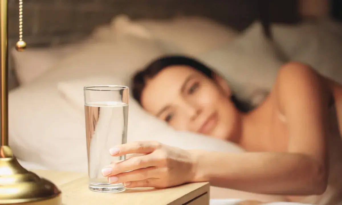 Une main attrape un verre d'eau posé à droite sur une table de chevet, une femme se réveille en arrière-plan.