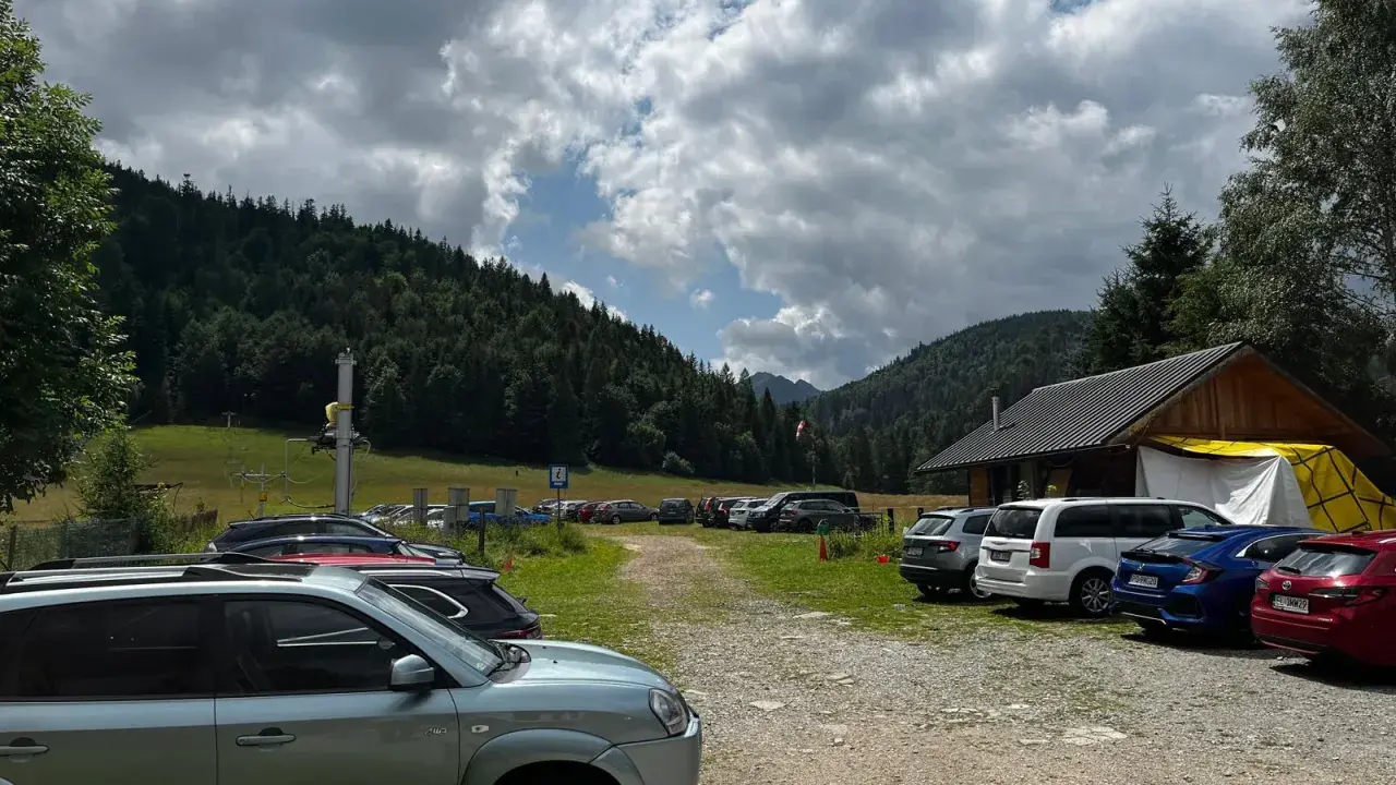 Parking z samochodami przy górskim schronisku, w tle las i chmury.
