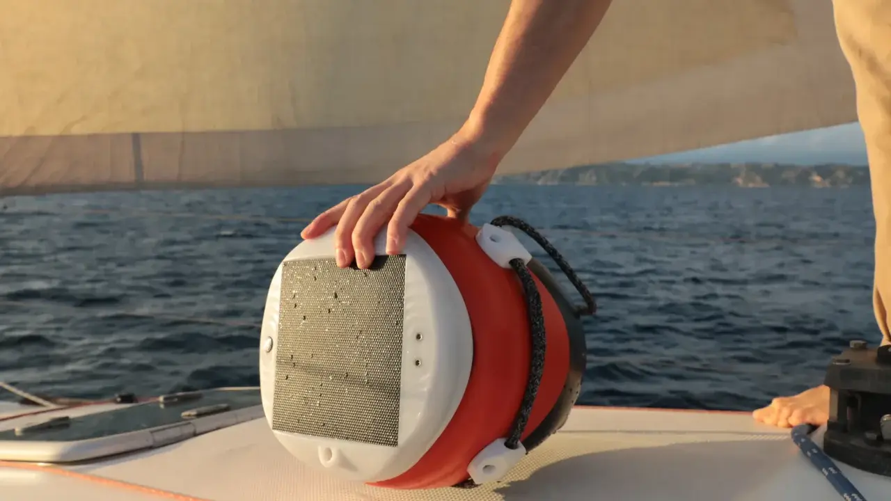 Main d'un marin tenant une bouée orange et blanche, un câble y est accroché. Le bateau navigue sur une mer bleue sous un ciel clair.
