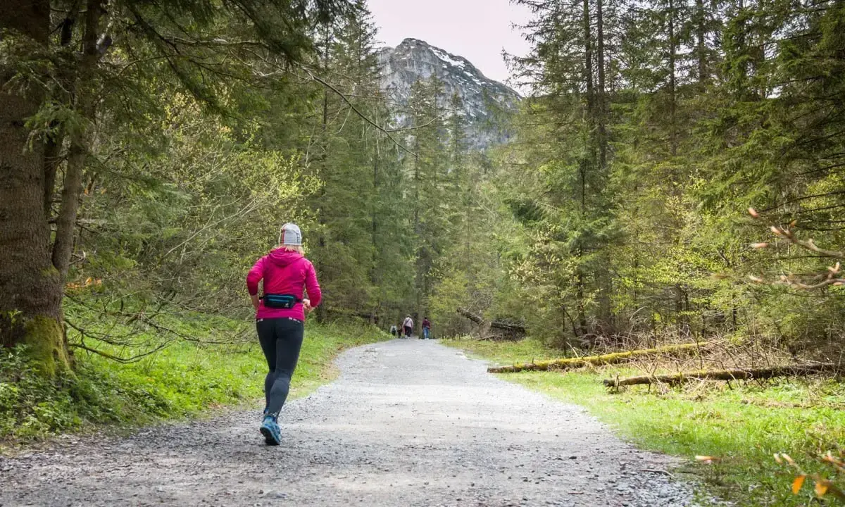 Kobieta w różowej bluzie biegnie leśną ścieżką, ciesząc się bieganiem po górach. W tle widać szczyt góry i innych turystów.