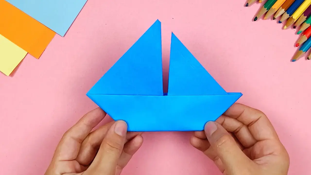 Hände falten ein blaues papierschiffchen. Bunte Stifte und Papier liegen bereit, um das papierschiffchen bauen zu beginnen.