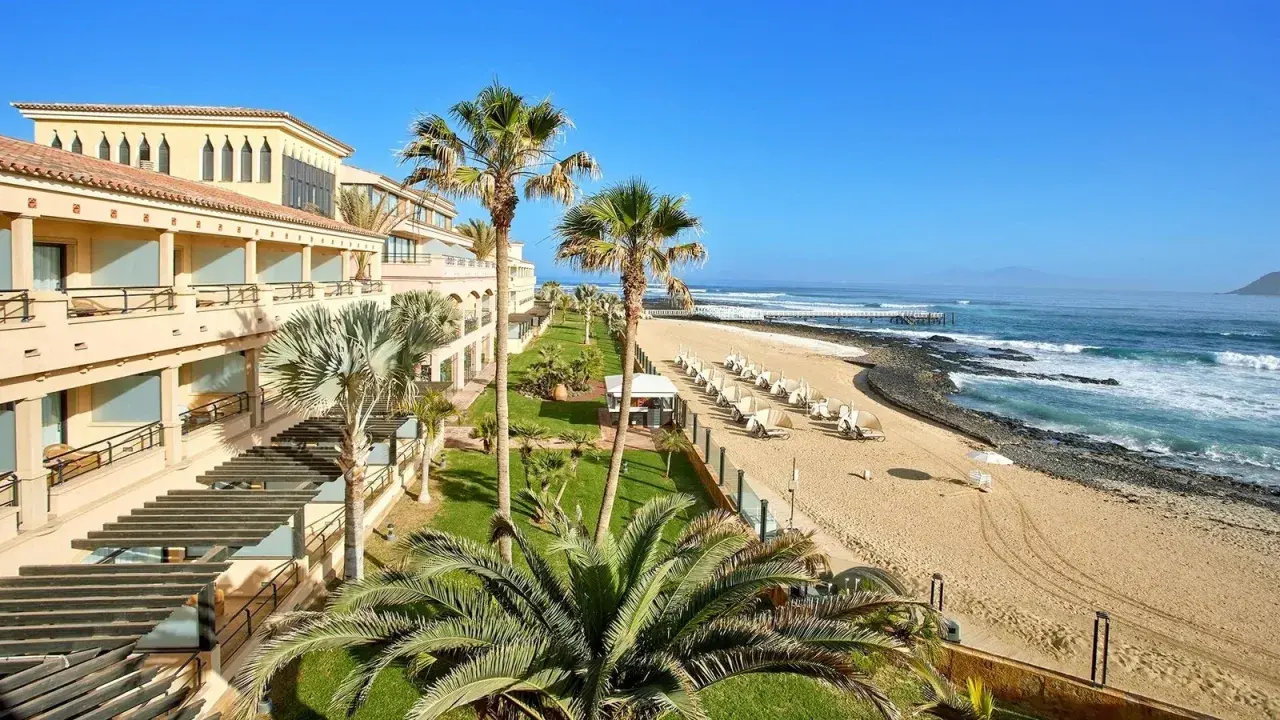 Fuerteventura kiedy wieje, palmy kołyszą się nad piaszczystą plażą i oceanem. Hotel z tarasami i widokiem na fale.