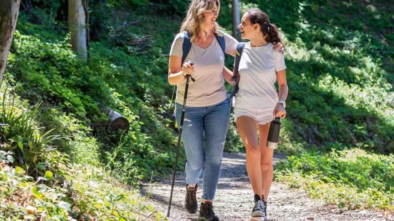 Två glada kvinnor njuter av en promenad i skogen, med en vandringsstav och en vattenflaska.