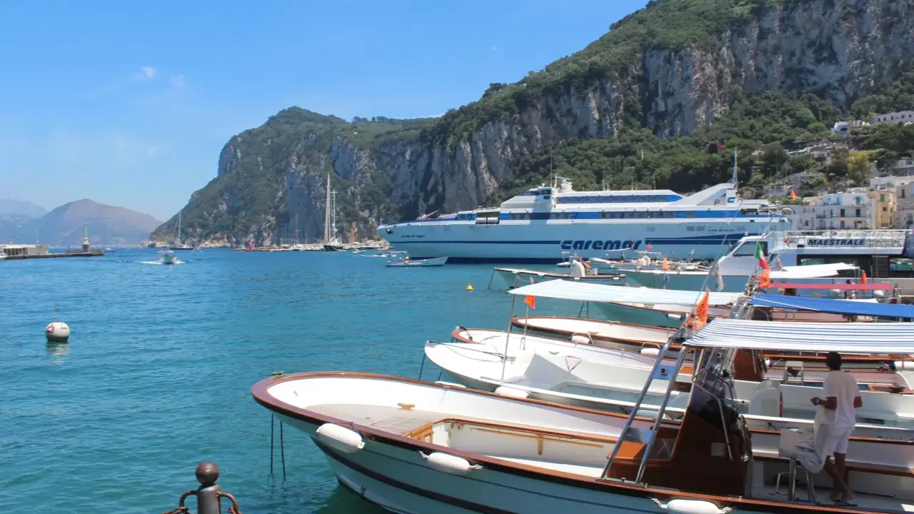 Barche in attesa al porto, con la maestosa isola di Capri sullo sfondo. Un traghetto Caremar attende passeggeri da Napoli.