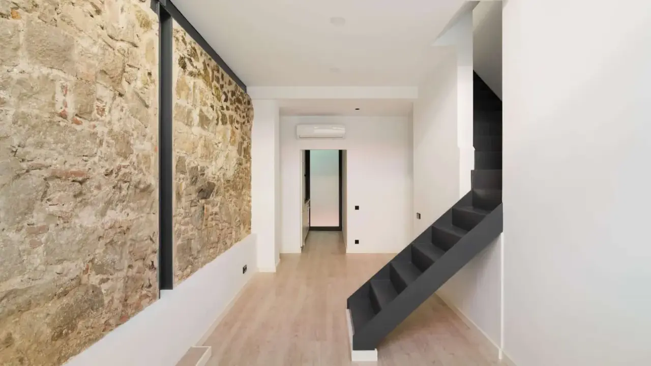 Interior moderno con pared de piedra rústica, escaleras oscuras y suelo de madera clara. Un ejemplo de cómo hacer una casa sostenible combinando lo antiguo y lo nuevo.