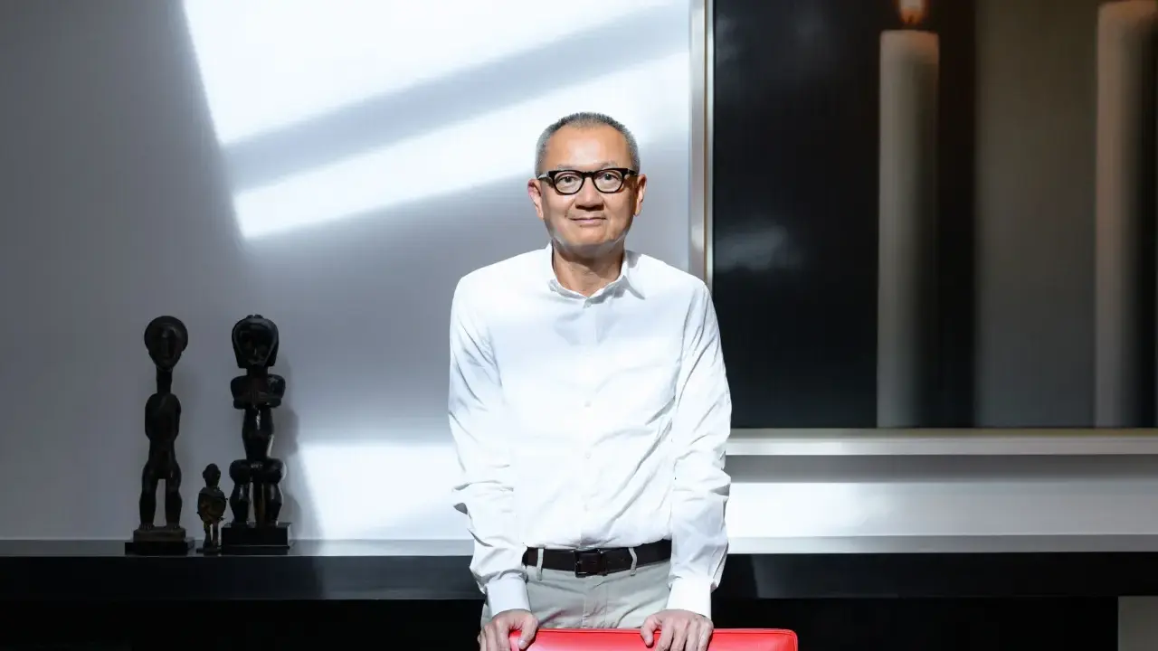 Pierre Chen, a man with glasses, stands in a modern room with striking shadows. He wears a white shirt and tan pants, leaning on a red chair.