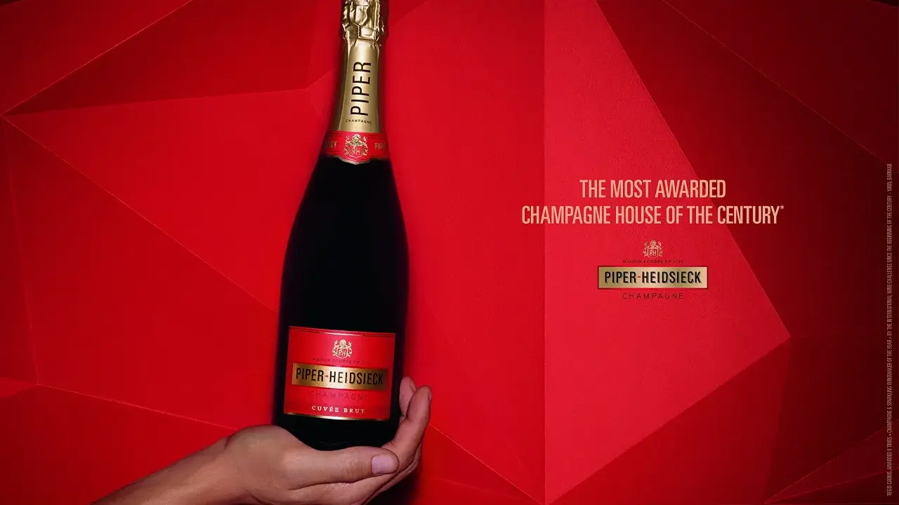 Eine Hand hält eine Flasche Piper-Heidsieck Champagner vor rotem, geometrischem Hintergrund.