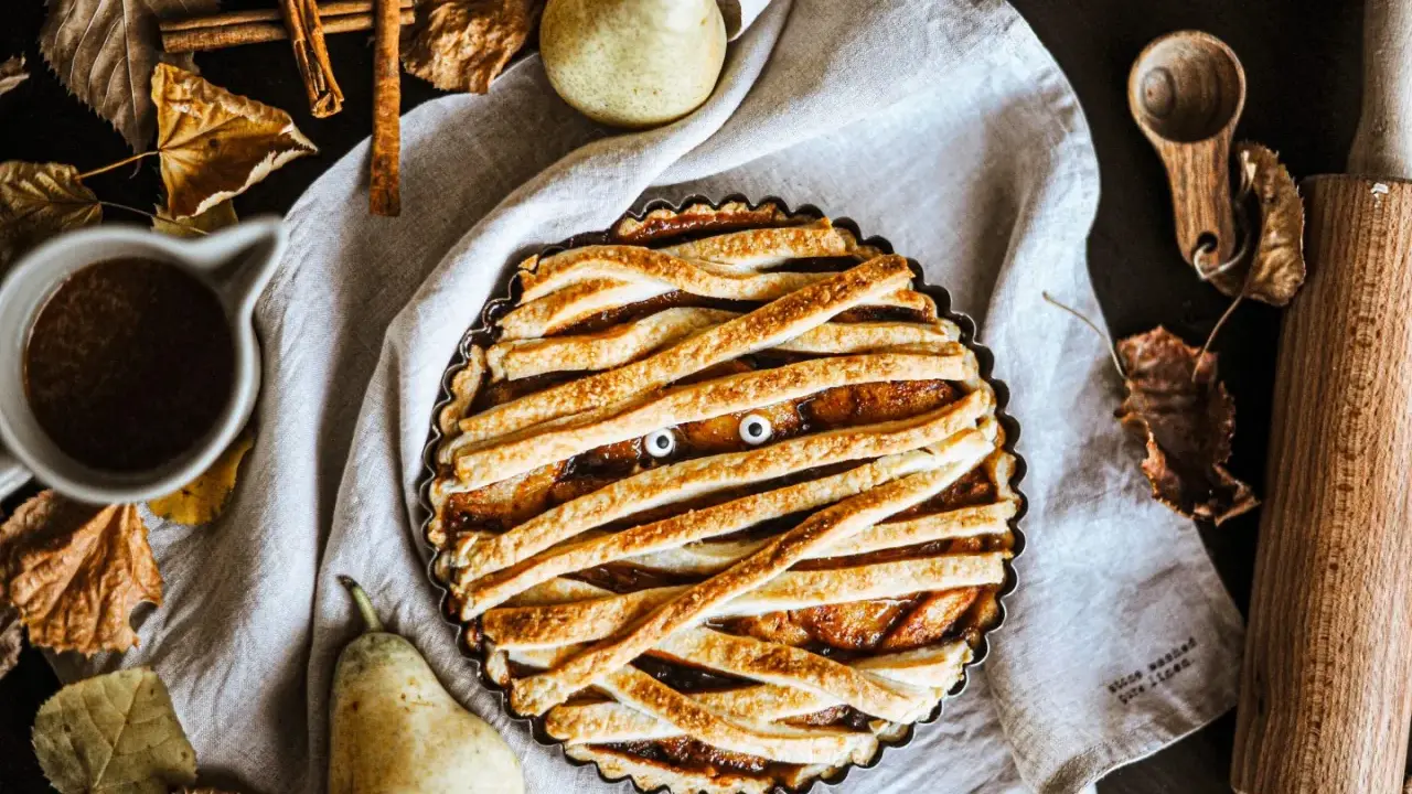 Tarte d'Halloween en forme de momie, avec des yeux globuleux et une garniture aux fruits. Autour, des feuilles d'automne, des poires et des épices.