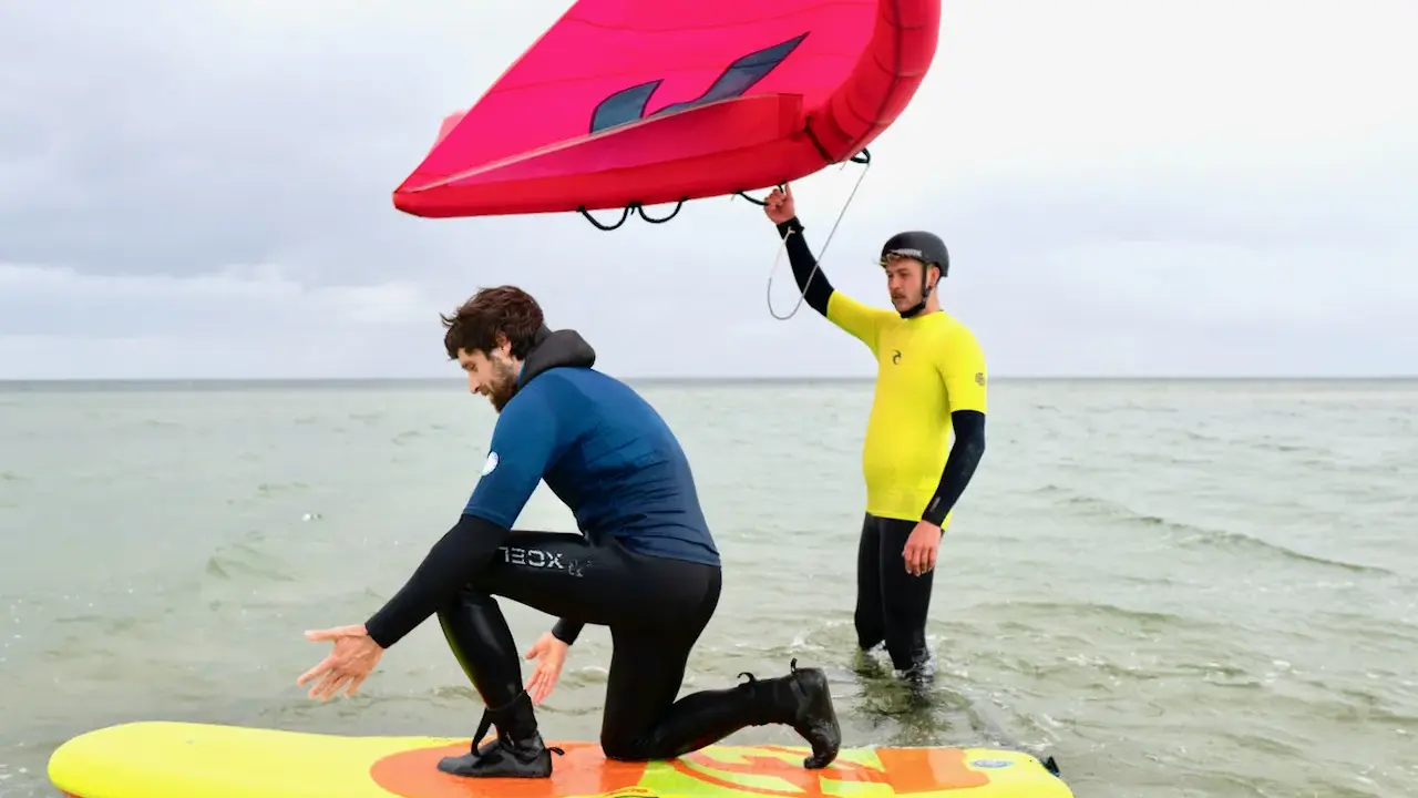 Zwei Männer üben Wing Surfen im flachen Wasser. Einer kniet auf einem gelben Board, der andere hält einen roten Wing.