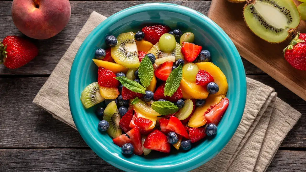 Un bol turquoise débordant de salade de fruits été : fraises, myrtilles, kiwis, pêches et raisins, garnie de menthe fraîche.