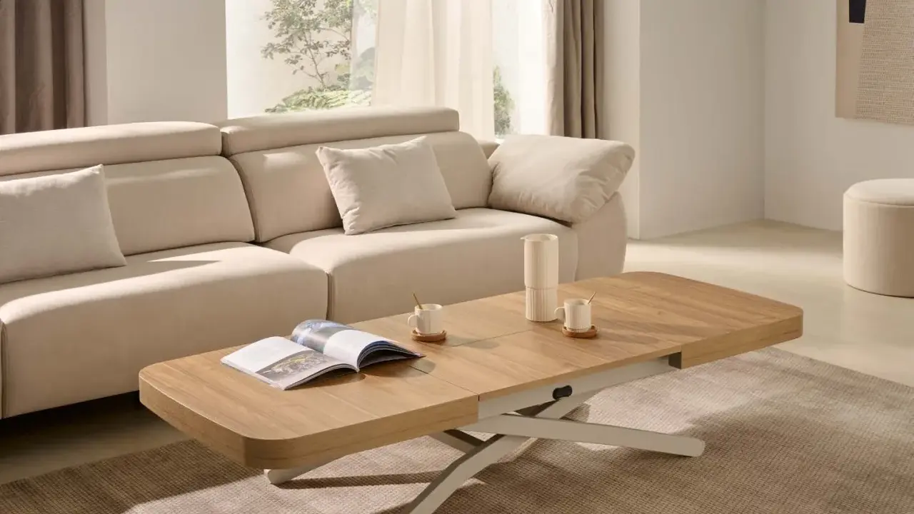 Mesa centro elevable 120x70 de madera clara, con un libro abierto y taza de café, junto a un sofá beige en un salón luminoso.