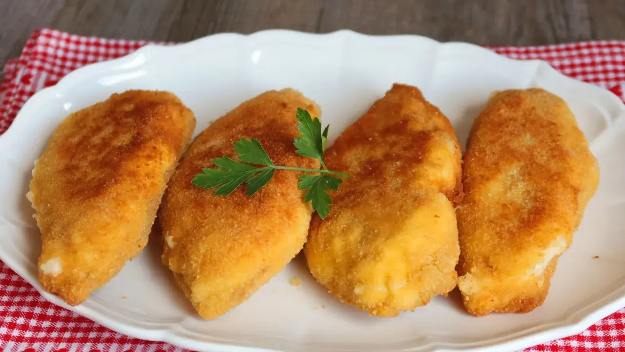 Cuatro pechugas villaroy doradas y crujientes, servidas en un plato blanco con una ramita de perejil.