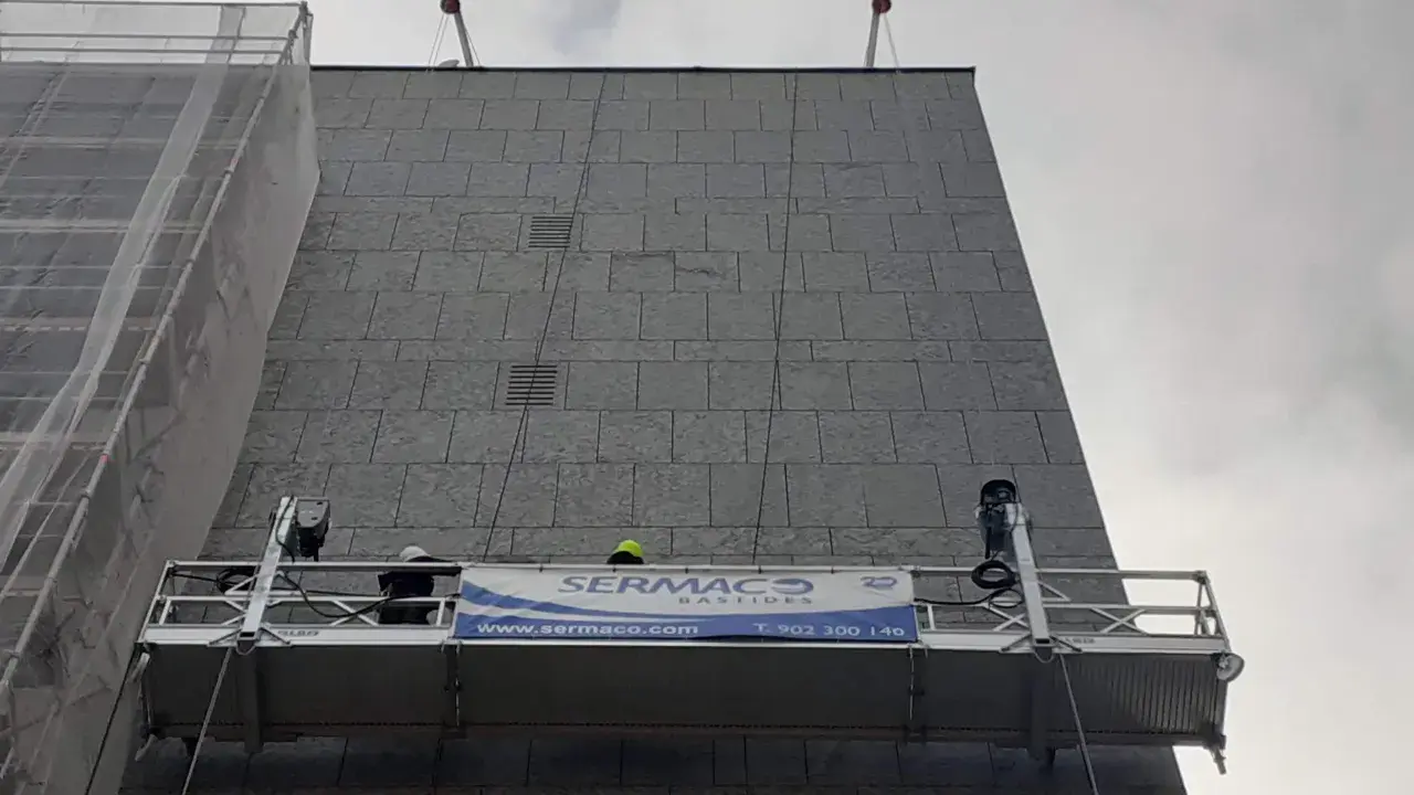 Dos trabajadores en andamios suspendidos realizan labores en la fachada de un edificio.