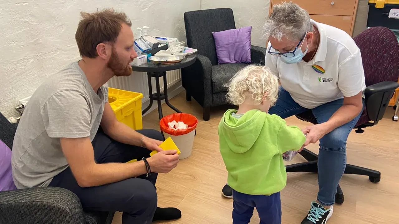 En pappa och ett barn får vaccin av en sjuksköterska. Barnet verkar lite oroligt, men sjuksköterskan är lugn och trygg. Det är en fin stund av vaccin italien.