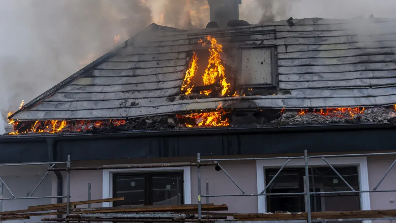 Un **incendie habitation** ravage le toit d'une maison, avec des flammes vives et une épaisse fumée s'échappant des combles.