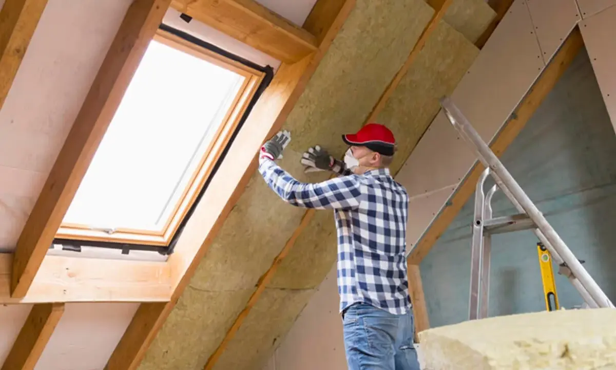 Mann isoliert ausgebauten Dachboden mit Dämmmaterial. Ein Dachfenster sorgt für Licht.