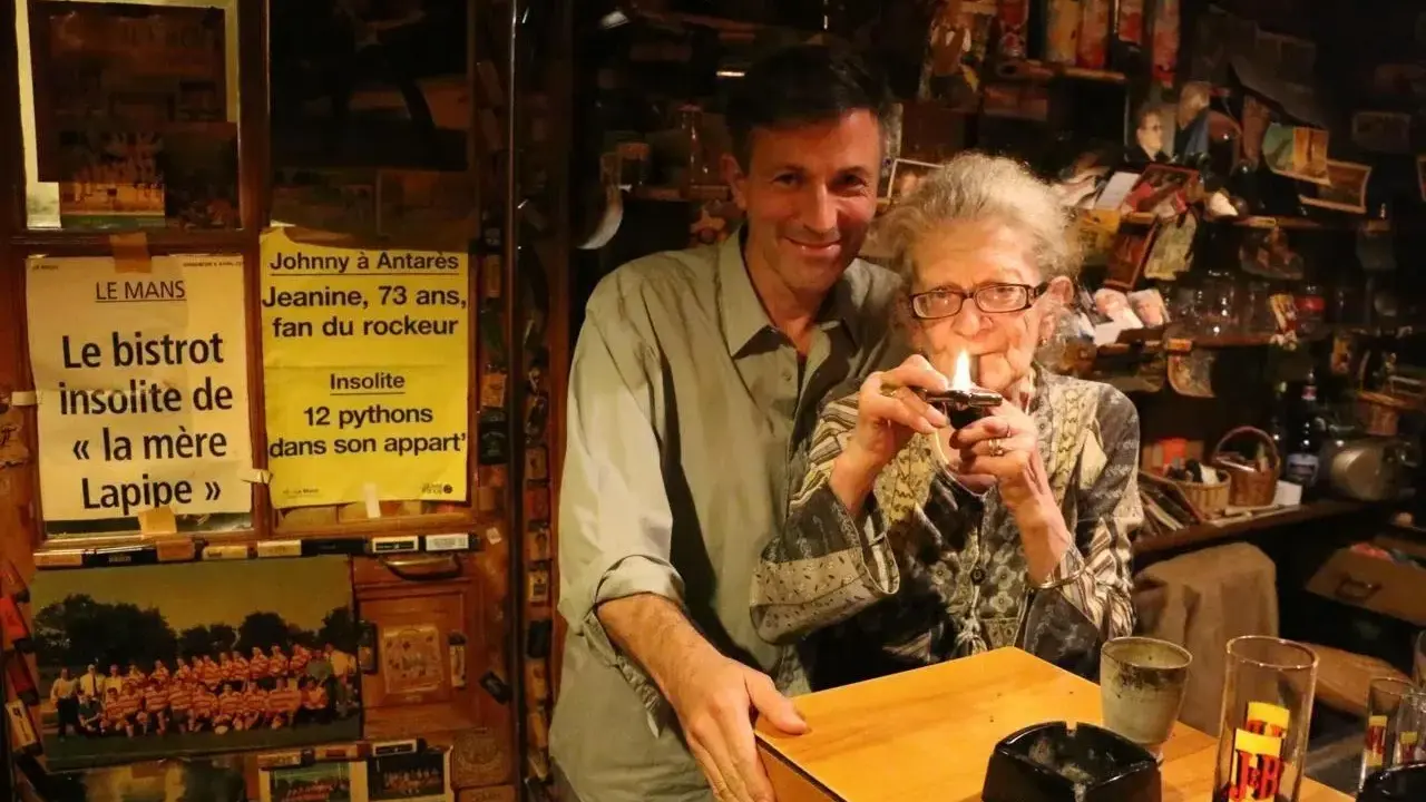 Au café du coin Le Mans, Jeanine, 73 ans, fan de rockeur, allume sa pipe. Un homme souriant se tient à ses côtés.