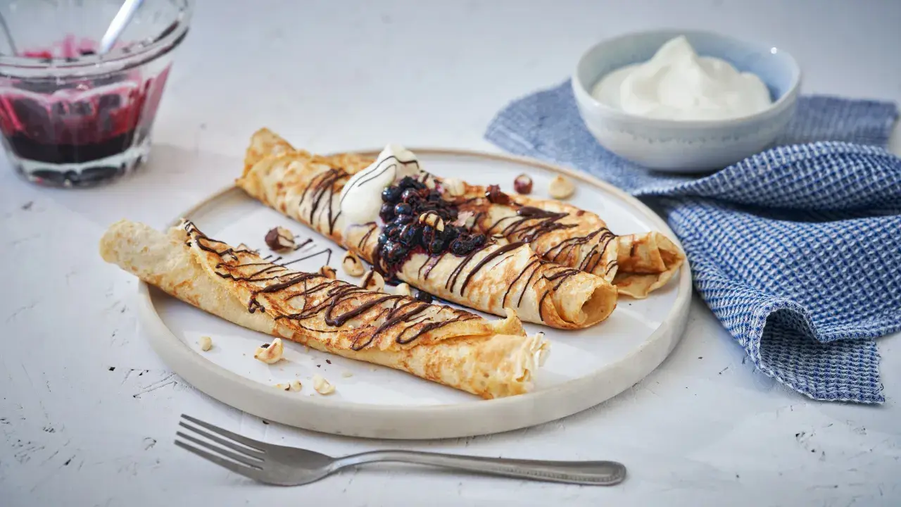 Matiga pannkakor rullade och toppade med chokladsås, blåbär och grädde. Serveras med en skål bärkompott och en blå rutig handduk.
