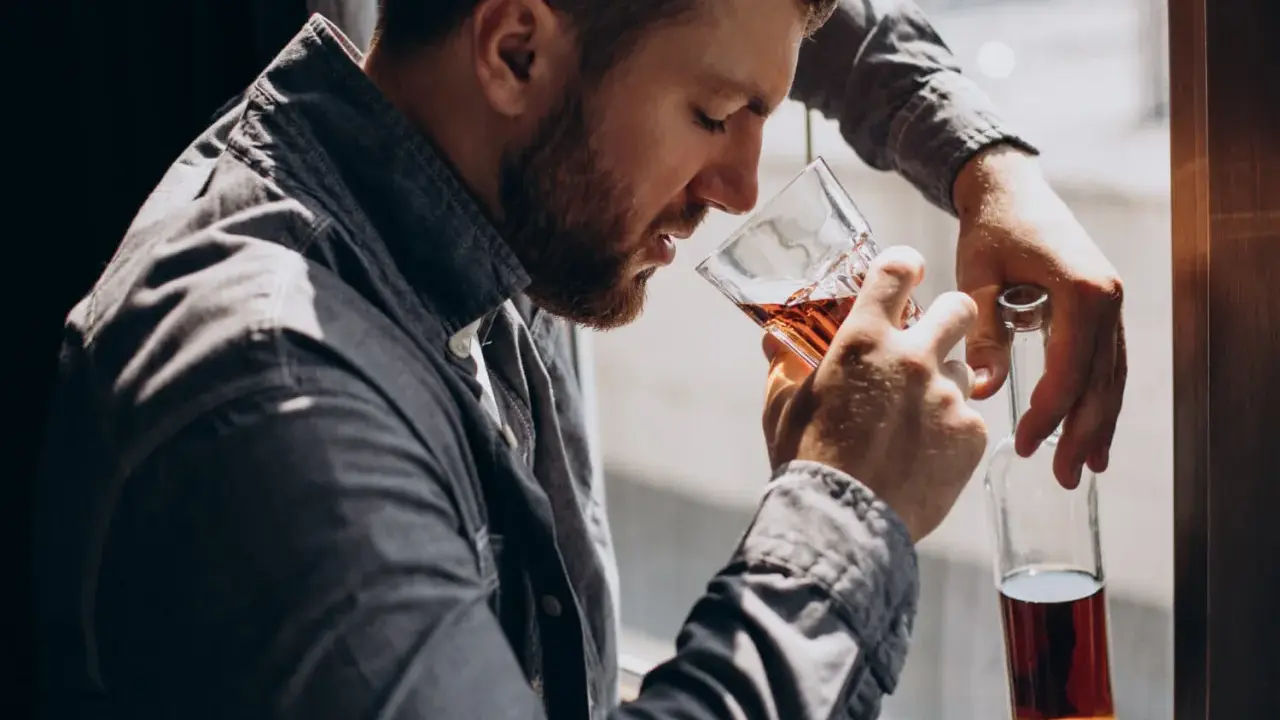 Mężczyzna z brodą, oparty o okno, pije alkohol. W ręku trzyma szklankę z bursztynowym płynem i pustą butelkę. To obraz, jak alkoholik zapije się na śmierć.