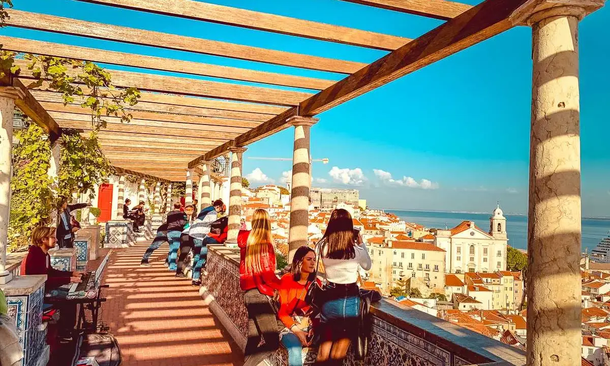 Blick vom Miradouro Lissabon über die Stadt und das Meer. Menschen genießen die Aussicht unter einer hölzernen Pergola.