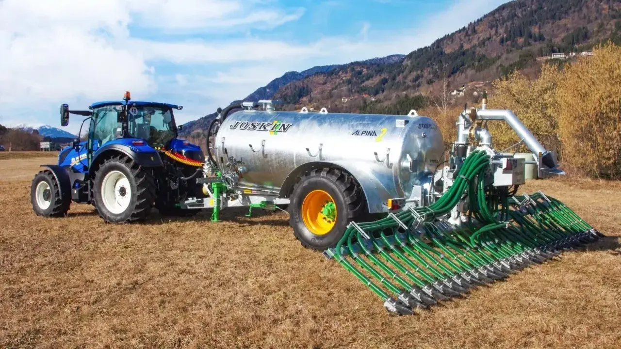 Niebieski traktor ciągnie przyczepę z cysterną do przewozu gnojowicy, wyposażoną w rozdzielacz z wieloma wężami do precyzyjnego nawożenia pól.