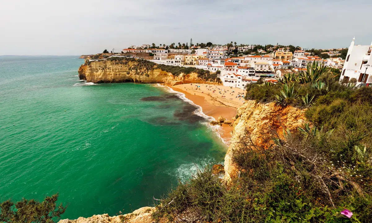 Blick auf die sonnenverwöhnte **Carvoeiro Altstadt** mit ihren weißen Häusern, die sich an die Klippen schmiegen. Der goldene Sandstrand lädt zum Verweilen ein, während das smaragdgrüne Meer sanft an die Küste brandet.