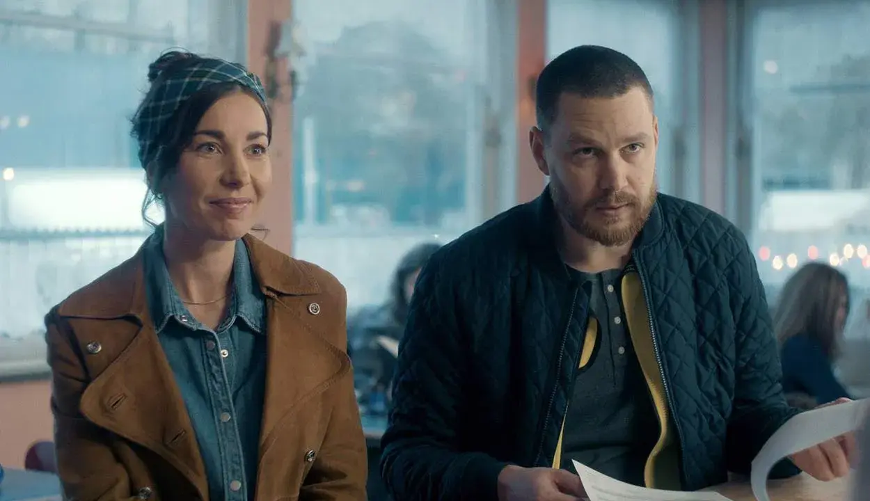 En kvinna med scarf och en man med skägg sitter vid ett bord. De ser ut att vara med i en dansk serie på SVT.