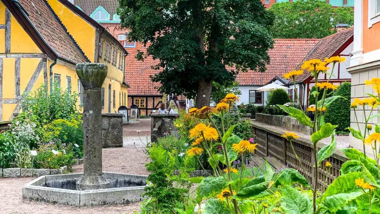 En vacker innergård i Lund utomhus med gula hus, en gammal fontän och blomstrande växter.