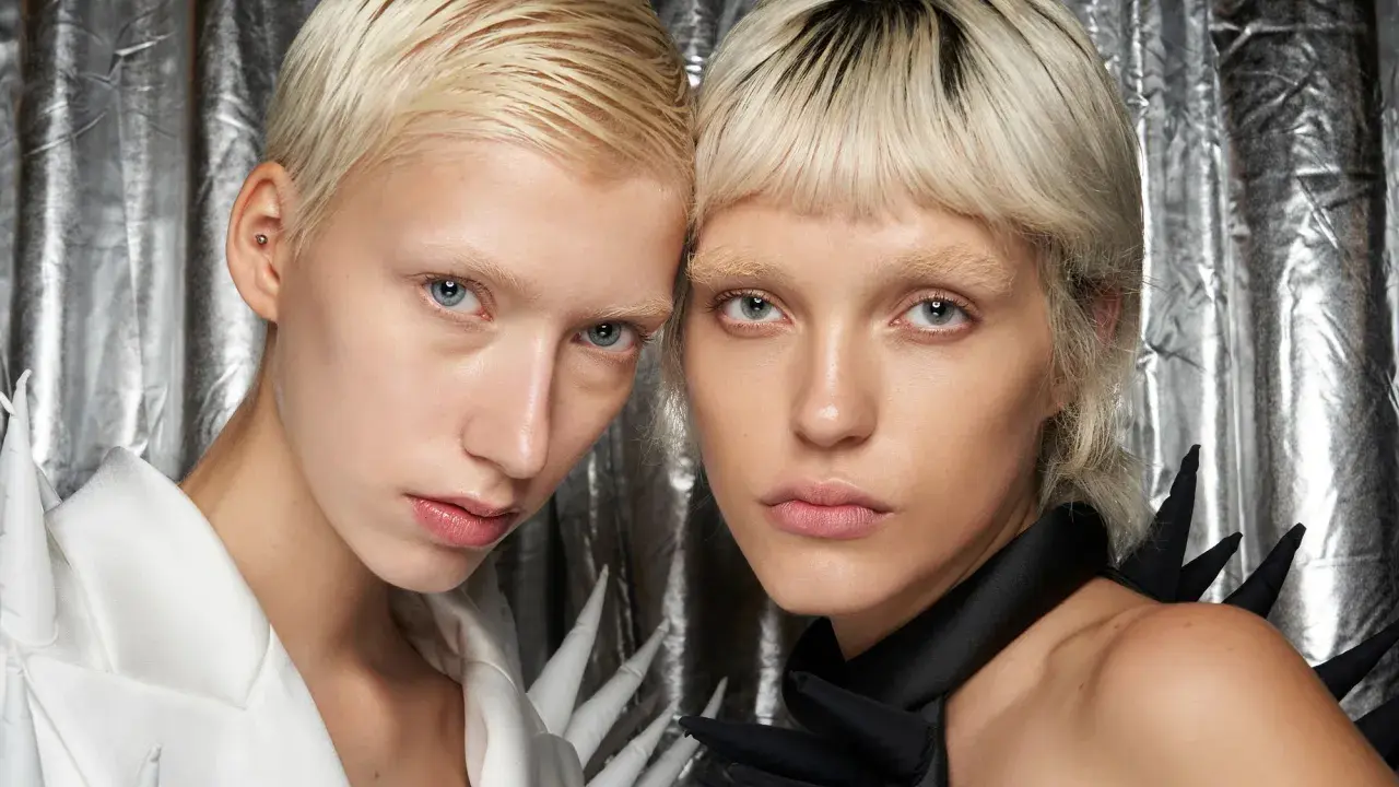 Zwei Models mit Pixie Cut feinem Haar und auffälligen Outfits vor silberner Kulisse.