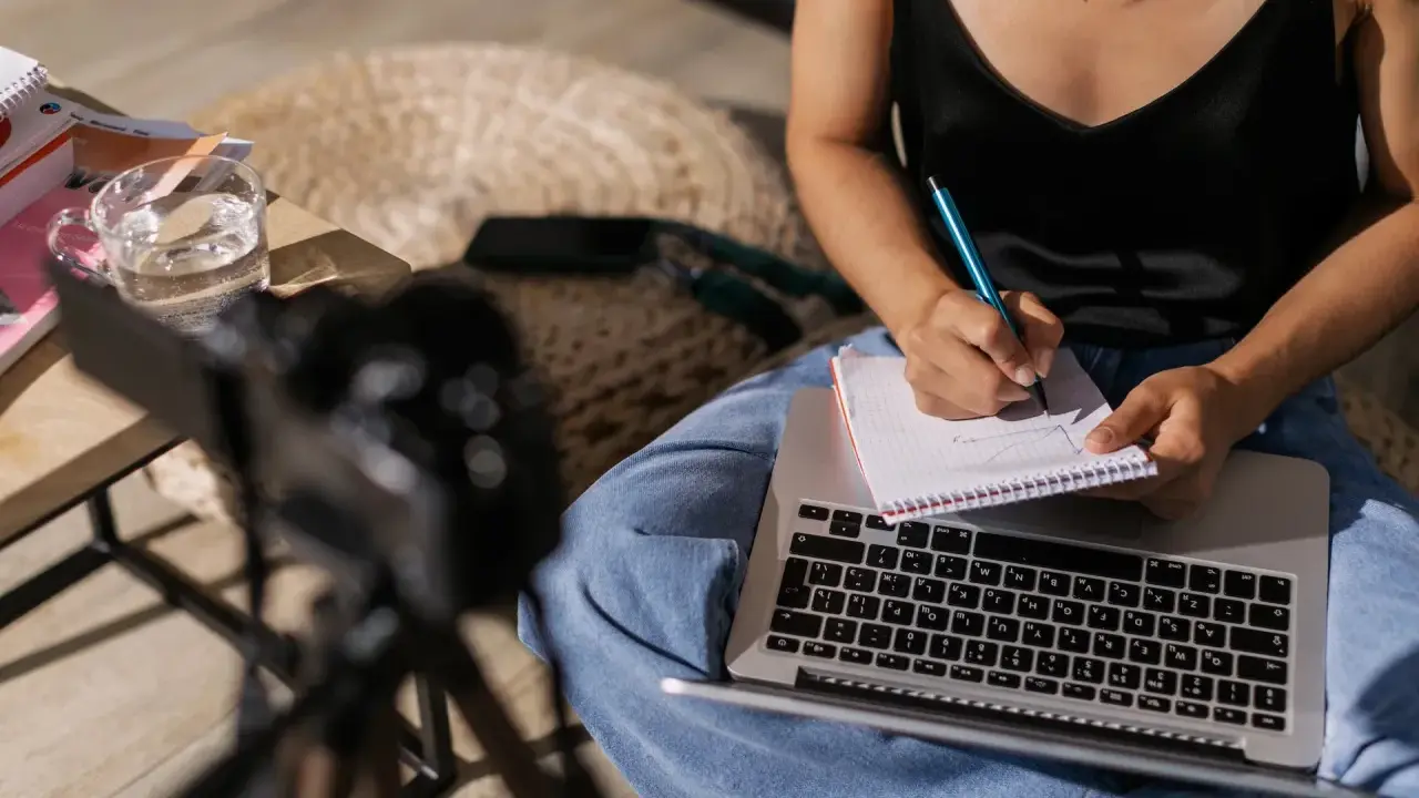 Kobieta pisze notatki w zeszycie, przygotowując materiał na artykuł. Obok leży laptop i szklanka wody.