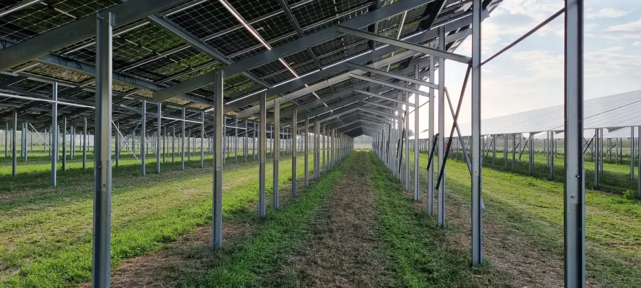 Reihe von Metallpfosten trägt eine agri-pv unterkonstruktion für Solarmodule. Die Struktur ist auf einem Feld mit Gras und Unkraut aufgebaut.