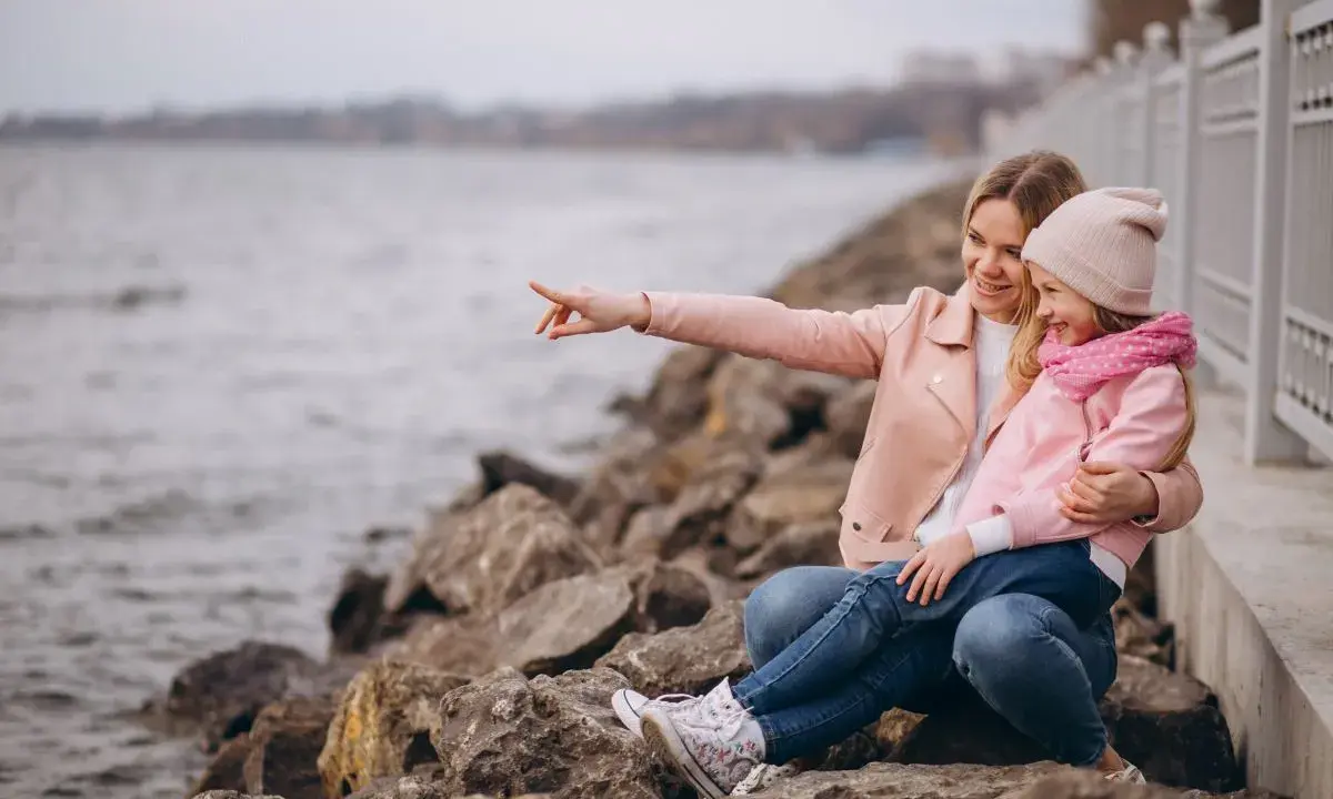 Mama z córką siedzą na kamieniach nad jeziorem. Mama wskazuje coś palcem, a dziewczynka się śmieje. To świetne gąski atrakcje dla dzieci.