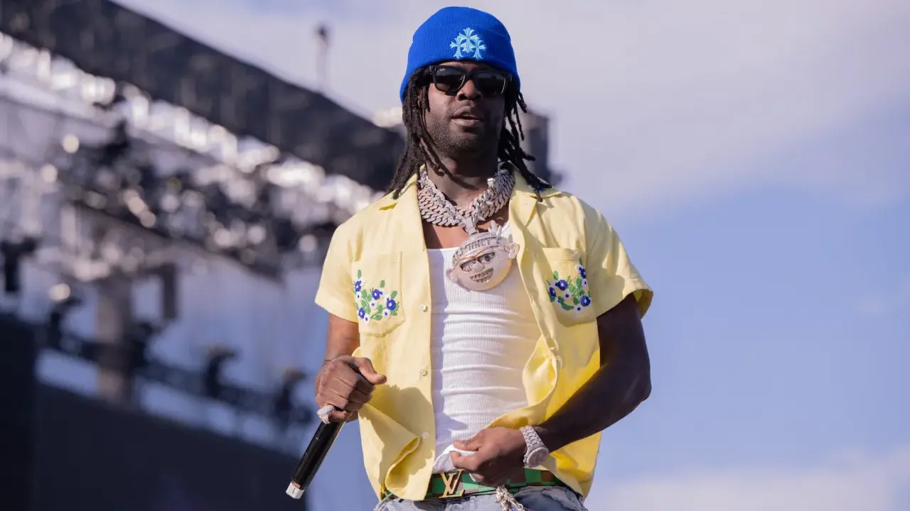 Chief Keef, w niebieskiej czapce i żółtej koszuli z kwiatami, na scenie. Jego wiek nie jest widoczny, ale styl mówi wszystko.