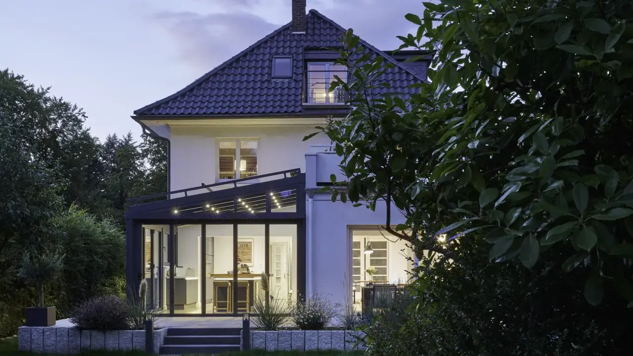 Moderner Anbau Wintergarten mit Glasfront und Beleuchtung erweitert ein Haus. Der Garten ist abends beleuchtet.