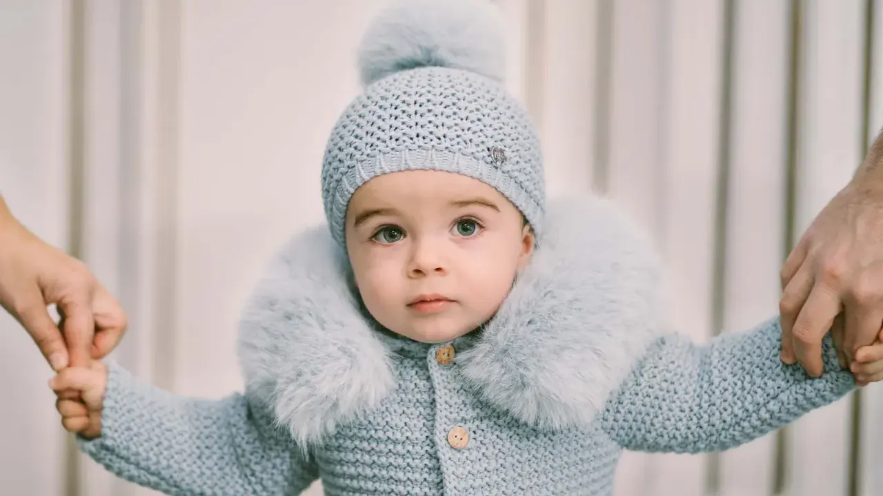 Un tierno bebé con un conjunto de punto azul claro, gorro y abrigo con cuello de pelo. Sus manos son sujetadas por adultos, como si estuviera aprendiendo a caminar. Este patrón de pelele para bebé es adorable.