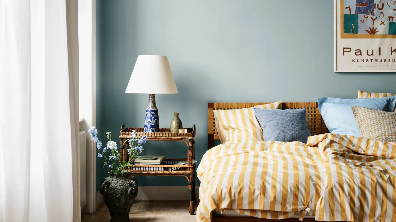 Dormitorio con paredes azul claro y ropa de cama a rayas amarillas y blancas. Los relajantes colores para dormitorios se complementan con una mesita de noche de mimbre y un jarrón con flores.