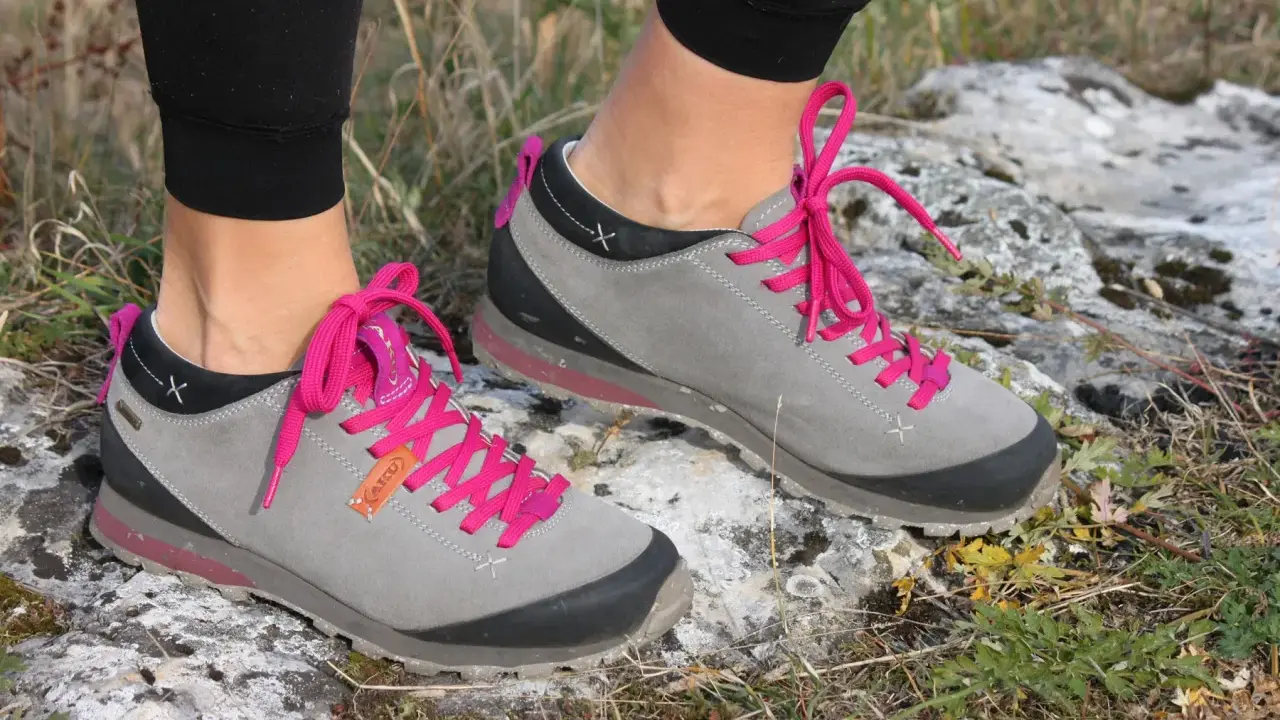 Szare buty trekkingowe Aku Bellamont z jaskrawymi, różowymi sznurówkami na kamienistym, trawiastym podłożu.