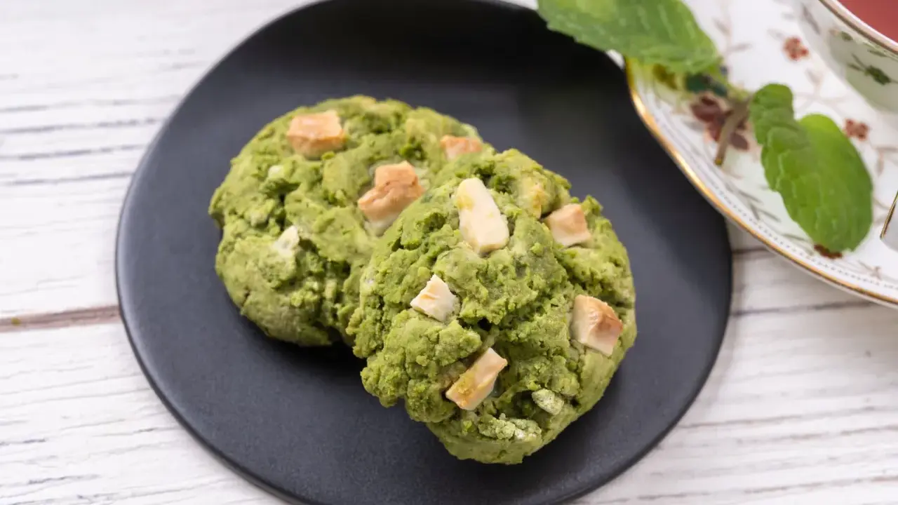 Deux cookies clairs au matcha, parsemés de morceaux de chocolat blanc, sur une assiette noire, à côté d'une tasse de thé et de feuilles de menthe.