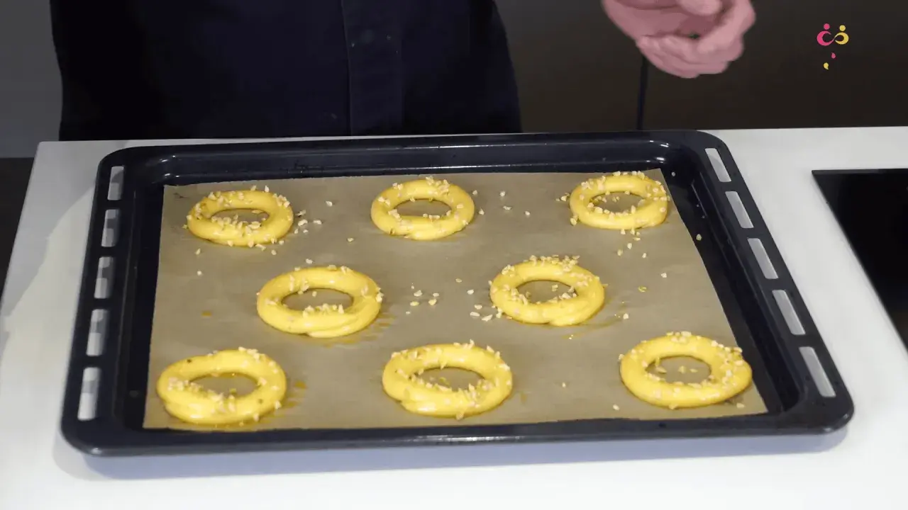 Huit ronds de pâte à choux, prêts à devenir un délicieux Paris-Brest, sont saupoudrés d'amandes effilées sur une plaque de cuisson.