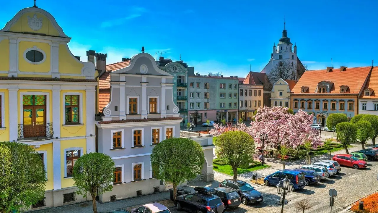 Kolorowy rynek w Kluczborku z zabytkowymi kamieniczkami, kwitnącą magnolią i wieżą kościelną. Kluczbork atrakcje zachwycają.