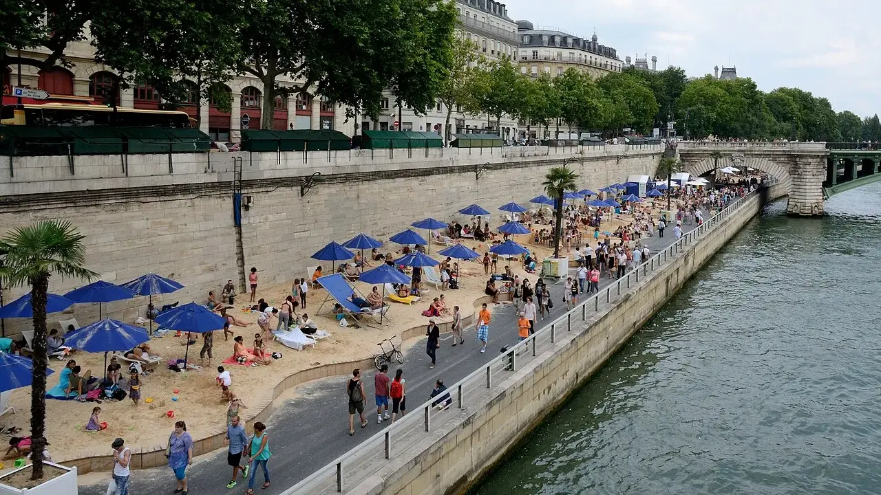 Menschen entspannen sich am **Paris strand** unter blauen Sonnenschirmen. Palmen und Gebäude säumen die Uferpromenade.
